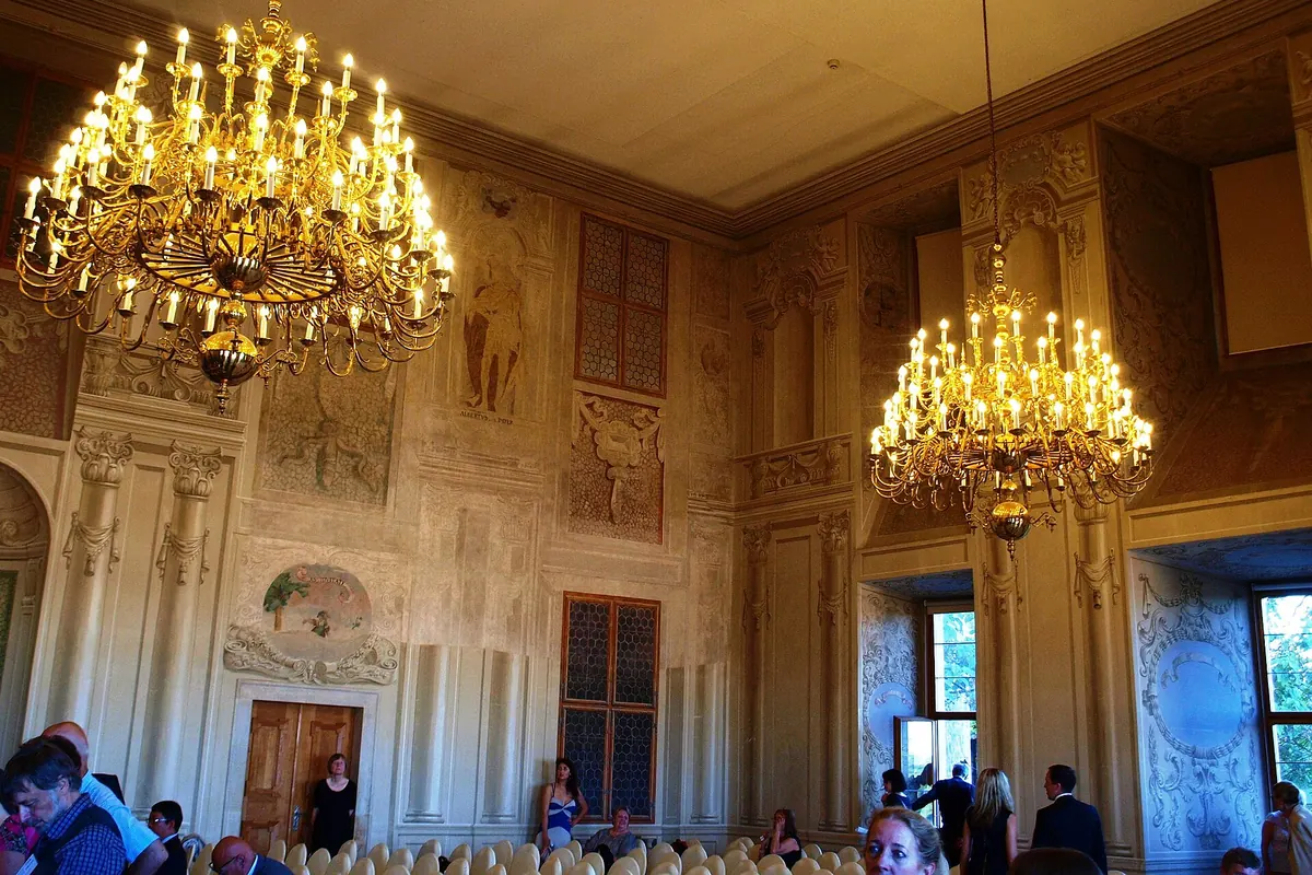 A hall of the palace with gilded chandeliers, painted walls and a window onto greenery.